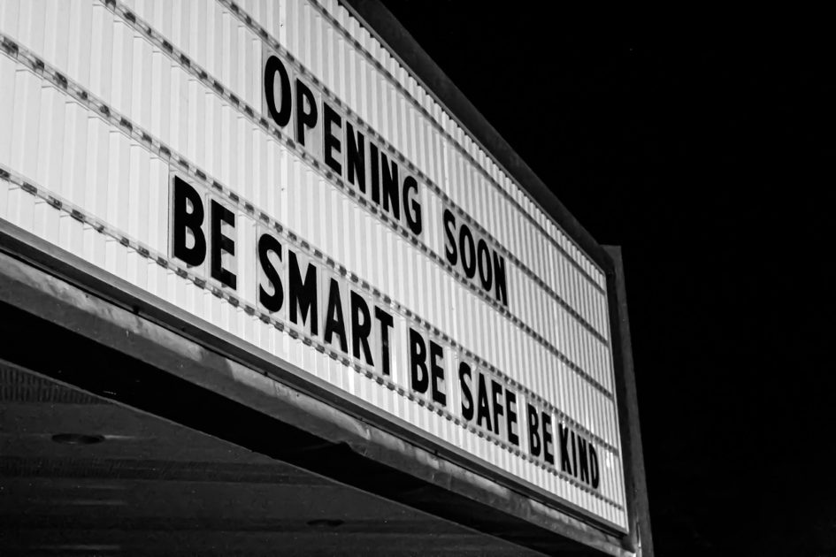 Eine große Texttafel überhalb eines Kino- oder Theatereingangs. Mit schwarzen Großbuchstaben steht dotr auf weißem Grund: Opening soo. Be smart. Be safe. Be kind.