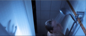 Frau unter der Dusche, die ihr Gesicht gerade leicht nach oben, dem Duschstrahl entgegen richtet. Links am Duschvorhang ist der Schatten einer Hand zu sehen.