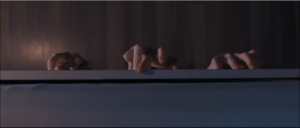 Die Kamera ist vom Bett aus nach unten auf den Parkettboden gerichtet. Drei Hände bewegen sich gerade unter dem Bett hervor und tasten teils am Bettkasten.