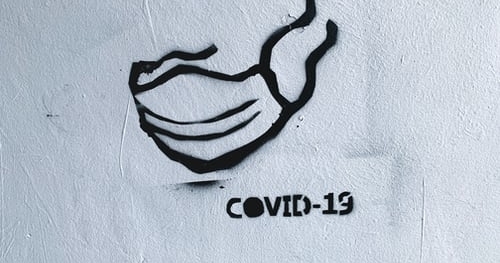 Ein Graffiti: mit schwarzer Farbe wurde ein Mund-Nasen-Schutz mit flatternden Bändern auf eine weiße Wand gesprüht. Darunter der Aufdruck Covid-19.