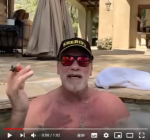 Zu sehen ist der Screenshot eines Youtube-Videos. Arnold Schwarzenegger sitzt oberkörperfrei, mit einer schwarzen Cap, einer Sonnenbrille und einer Zigarre in der rechten Hand in seinem Pool und spricht in die Kamera. Im Hintergrund abgedeckte Terrassenmöbel und eine sandfarbene Hauswand.