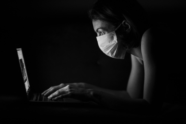 Eine Frau sitzt mit Mund-Nasen-Schutz vor ihrem aufgeklappten Laptop, sie lehnt sich vor in Richtung Bildschirm und tippt gerade etwas. Die Frau ist im Profil zu sehen und wird nur durch den Bildschirm beleuchtet, der Hintergrund ist schwarz. Der Mund-Nasen-Schutz der Frau leuchtet weiß und sticht dadurch hervor.