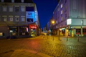 Der nächtliche, menschenleere Hans-Albers-Platz in Hamburg, St. Pauli. Verlassene Bars und Kneipen, davor eine Straße mit Kopfsteinpflaster. Einzelne Lichtquellen sind die teilweise rot erleuchteten Schriftzüge der Barnamen und eine orangene Straßenlaterne.