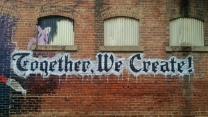 Ein Graffitti an einer roten Backsteinmauerwand. Der Schriftzug heißt: Together we create. Die Buchstaben sind schwarz, haben aber eine weiße Umrandung.